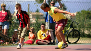Two boys play football on an outdoor court at Camping Bella Italia while people watch in the background.