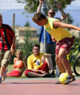 Dos chicos juegan fútbol en una cancha al aire libre en Camping Bella Italia con espectadores detrás.