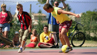 Dos chicos juegan fútbol en una cancha al aire libre en Camping Bella Italia con espectadores detrás.