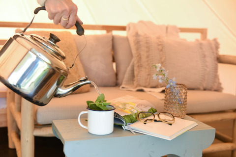 Een hand giet thee uit een ketel in een tas op een tafel met bril, boek en vaasje in een safari-tent.