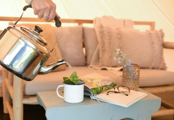 Een hand schenkt thee uit een ketel in een mok op een tafel met boek, bril en vaas in een safari-tent.