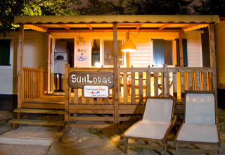 Vista nocturna de la cabaña SunLodge Maple en Camping Bella Italia, Italia, con terraza iluminada y tumbonas.