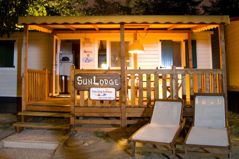 Vista nocturna de la cabaña SunLodge Maple en Camping Bella Italia, Italia, con terraza iluminada y tumbonas.
