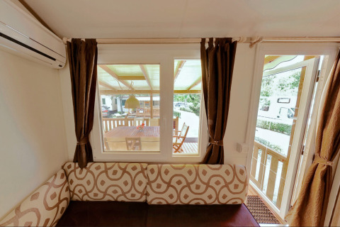 Vista interior de SunLodge Maple en Camping Bella Italia, con sofá, cortinas y zona de terraza exterior.