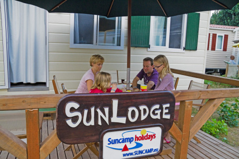 Famiglia si rilassa sulla terrazza della SunLodge Maple al Camping Bella Italia, Italia, sotto un ombrellone.
