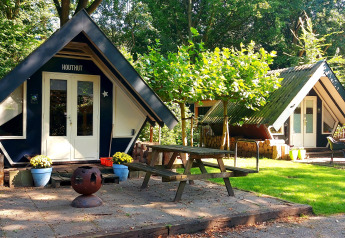 Billede af to små Trekkershut-hytter i en grøn, solrig skov med picnicbord og ildsted ude foran.