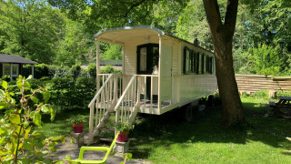 Das Tiny House Pipowagen steht zwischen Bäumen im Grünen mit Terrasse und Gartenmöbeln im Sonnenschein.
