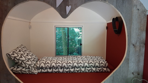 Acogedora habitación con abertura en forma de corazón, cama individual y ventana hacia árboles verdes.
