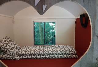 Acogedora habitación con abertura en forma de corazón, cama individual y ventana hacia árboles verdes.