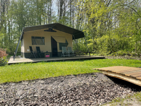 Tienda safari sobre una plataforma de madera en un bosque, con muebles de exterior y un pequeño puente.