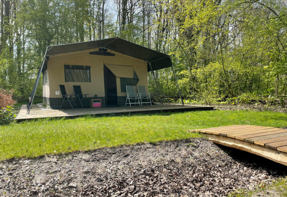 Tienda safari sobre una plataforma de madera en un bosque, con muebles de exterior y un pequeño puente.