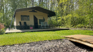 Tienda safari sobre una plataforma de madera en un bosque, con muebles de exterior y un pequeño puente.
