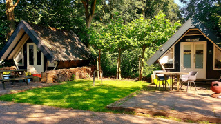 Dos pequeñas casas en el bosque, una con el cartel 'Trekkershut', rodeadas de mesas, sillas y césped.