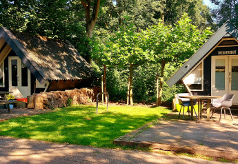 Dos pequeñas casas en el bosque, una con el cartel 'Trekkershut', rodeadas de mesas, sillas y césped.