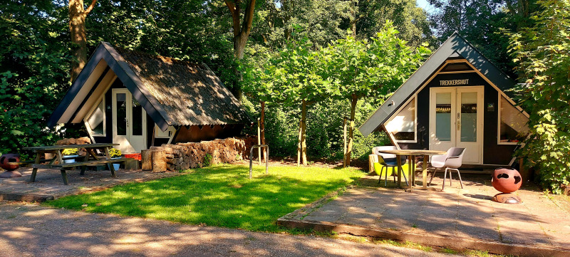 Dos pequeñas casas en el bosque, una con el cartel 'Trekkershut', rodeadas de mesas, sillas y césped.