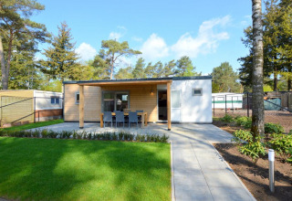 Moderne lodge med træfacade, terrasse og udendørs spiseplads, omkranset af grønt græs og skov.