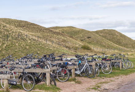 Bicicletas aparcadas en un parque vacacional con dunas cubiertas de hierba al fondo y cielo nublado.
