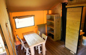 Interieur van een safaritent met houten tafel, stoelen, open kast en badkamer, in warm licht.
