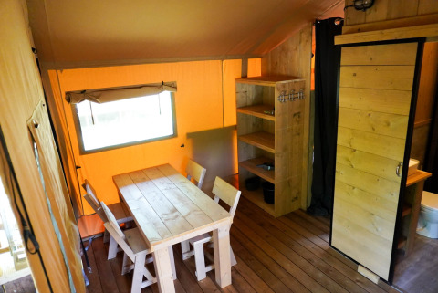 Interior de una tienda safari con mesa de madera, sillas, estantería y baño, iluminado con luz cálida.