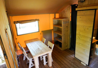 Intérieur d'une tente safari avec table en bois, chaises, étagère et salle de bain, éclairé chaleureusement.
