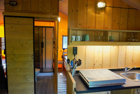 Intérieur d'une tente safari avec des panneaux en bois, coin cuisine, évier et salle de bain avec douche.