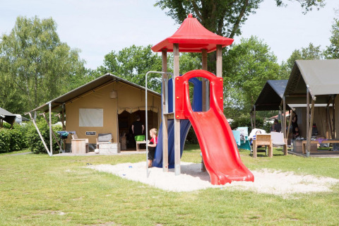 Niños jugando en un tobogán rojo junto a la tienda Comfort safari en Camping 't Veld, Países Bajos, rodeados de tiendas.