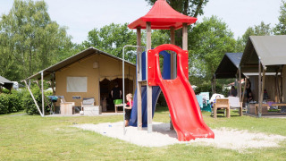 Niños jugando en un tobogán rojo junto a la tienda Comfort safari en Camping 't Veld, Países Bajos, rodeados de tiendas.