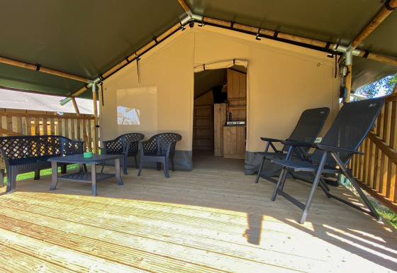 Tenda safari con veranda in legno, sedie di plastica e servizi igienici privati visibili all’interno.