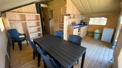 Interieur van een safaritent met grote tafel, zes stoelen, keukentje, rekken en koelkast.