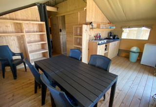 Vista interna di una tenda safari con tavolo da pranzo, sei sedie, angolo cottura, scaffali e frigo.