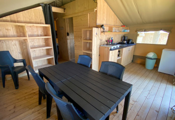 Interieur van een safaritent met grote tafel, zes stoelen, keukentje, rekken en koelkast.