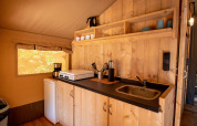 Kleine kitchenette in een safaritent met houten wanden, kookplaat, koelkastje en open planken.