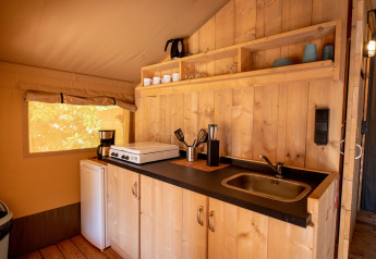 Compact kitchenette inside a safari tent, featuring wood paneling, mini fridge, sink, and open shelves.