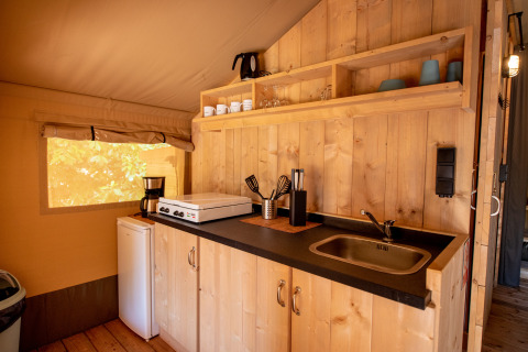 Pequeña cocina dentro de una tienda safari, con paredes de madera, mini nevera, fregadero y estantes.