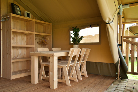 Interieur van een glamping tent met houten tafel en stoelen, boekenrek, plant en houten vloer.