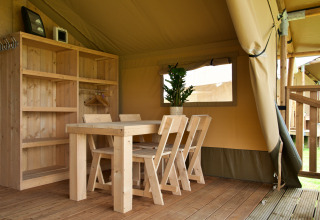 Intérieur d’une tente glamping avec table en bois, chaises, étagère, plante en pot et sol en bois.