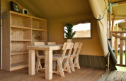 Intérieur d’une tente glamping avec table en bois, chaises, étagère, plante en pot et sol en bois.