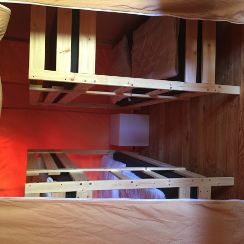 Interior de una tienda safari con dos literas de madera, ropa de cama blanca, almohadas y suelo de madera.