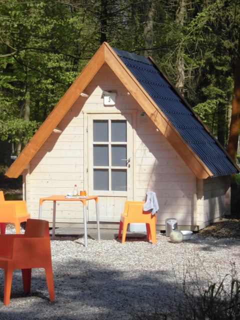 Pequeña cabaña de madera con sillas naranjas y mesa fuera, rodeada de bosque en Kabouterhuisje lodge.