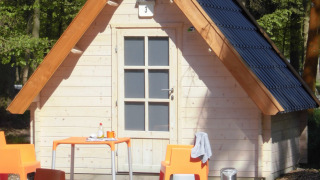 Piccola casetta di legno con sedie e tavolo arancioni fuori, immersa nel bosco al lodge Kabouterhuisje.