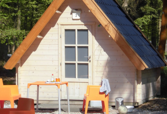 Kleine houten hut met oranje stoelen en tafel buiten, omgeven door bos aan Kabouterhuisje-lodge.