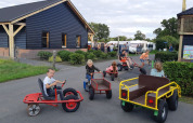 Børn leger på pedalbiler foran en bygning på Camping Nieuw Romalo i Gelderland, Holland, solrig dag.