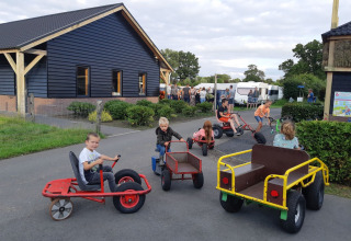 Børn leger på pedalbiler foran en bygning på Camping Nieuw Romalo i Gelderland, Holland, solrig dag.