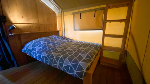 Interno di una tenda safari con letto matrimoniale, parete in legno, scala e luce gialla soffusa.
