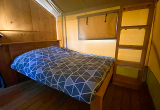Interior de una tienda safari con cama doble, pared de madera, escalera y cálida iluminación amarilla.