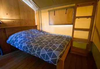 Interieur van een safaritent met een tweepersoonsbed, houten wand, ladder en warme gele verlichting.