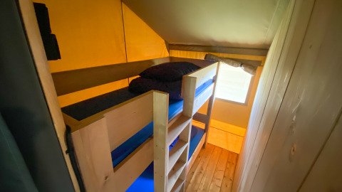 Vista interior de una tienda safari con literas de madera y ropa de cama azul junto a una pequeña ventana.