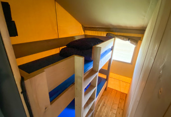 Vista interior de una tienda safari con literas de madera y ropa de cama azul junto a una pequeña ventana.