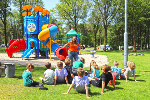 Kinder sitzen auf dem Rasen vor einem Spielplatz in Eurocamping Vessem, Niederlande, mit einer Betreuerin.