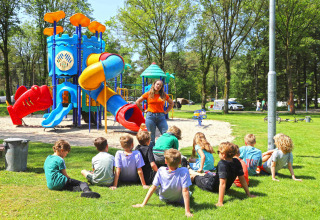 Kinder sitzen auf dem Rasen vor einem Spielplatz in Eurocamping Vessem, Niederlande, mit einer Betreuerin.